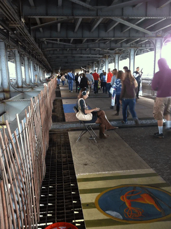 A woman wearing headphones sits beneath a steel bridge while festival visitors walk past.