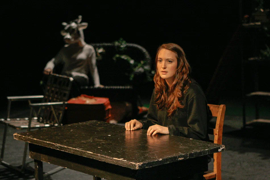 A woman sits at a table while a performer in a horned animal mask stands behind her.