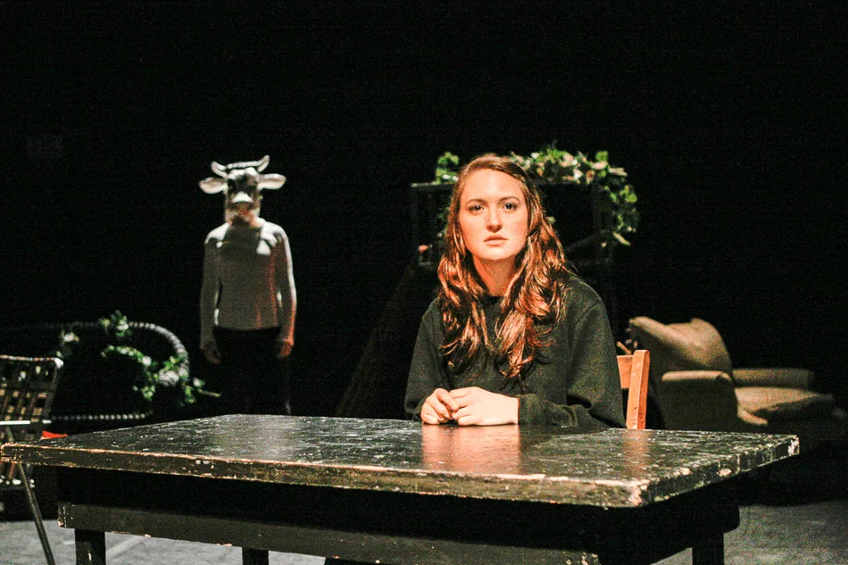 A woman faces forward at a table with a horned masked figure in the background.