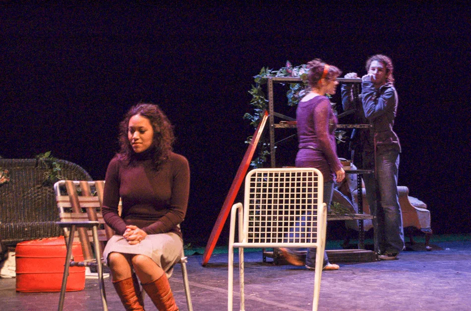 A seated woman faces the audience while two performers stand near a mirror behind her.