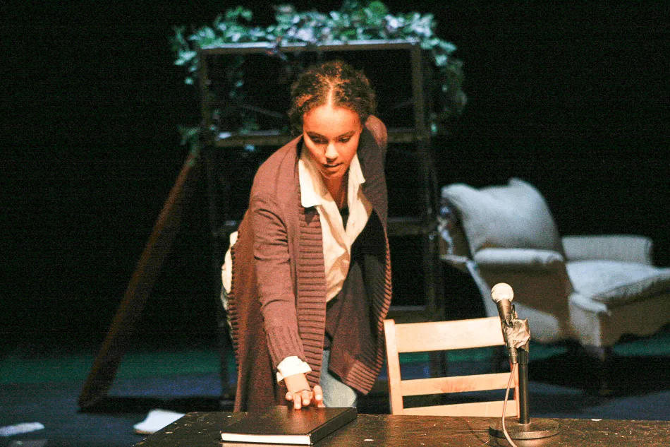 A woman in a blazer leans over a table on a sparsely furnished stage.