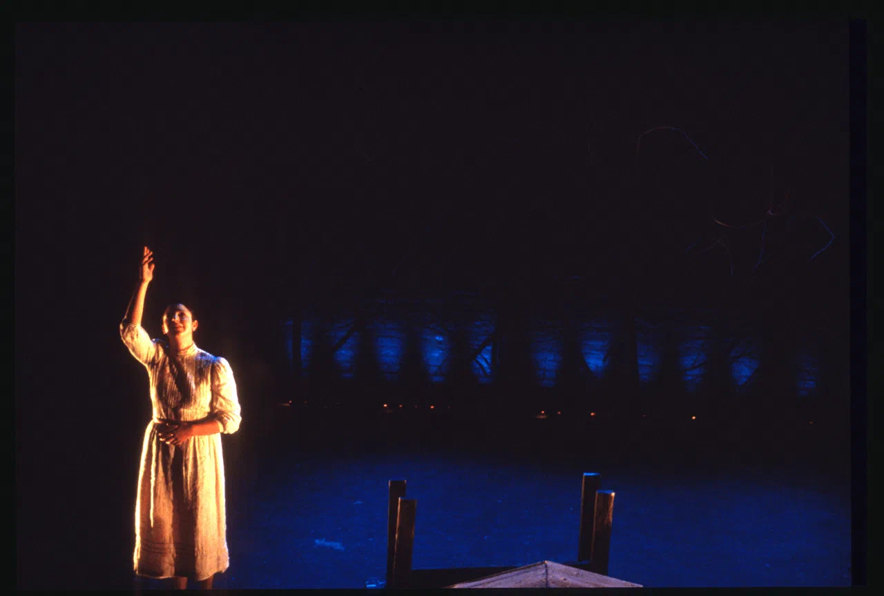 A woman in a long white dress raises one hand in a narrow pool of stage light.