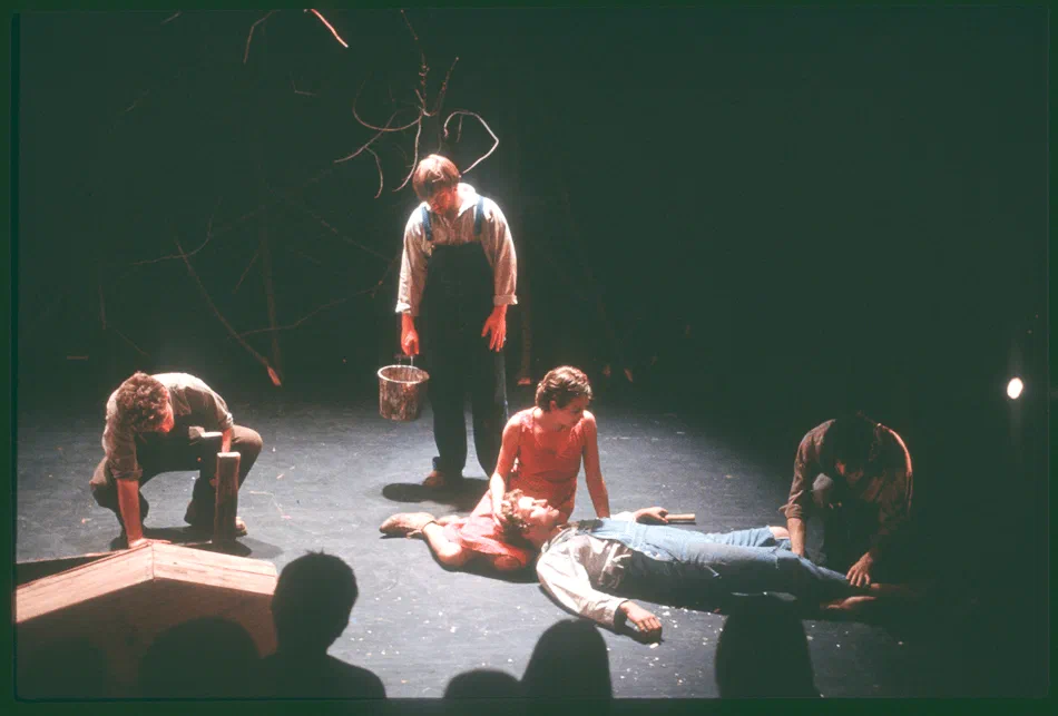 Performers surround a figure lying at the center of a dark stage.
