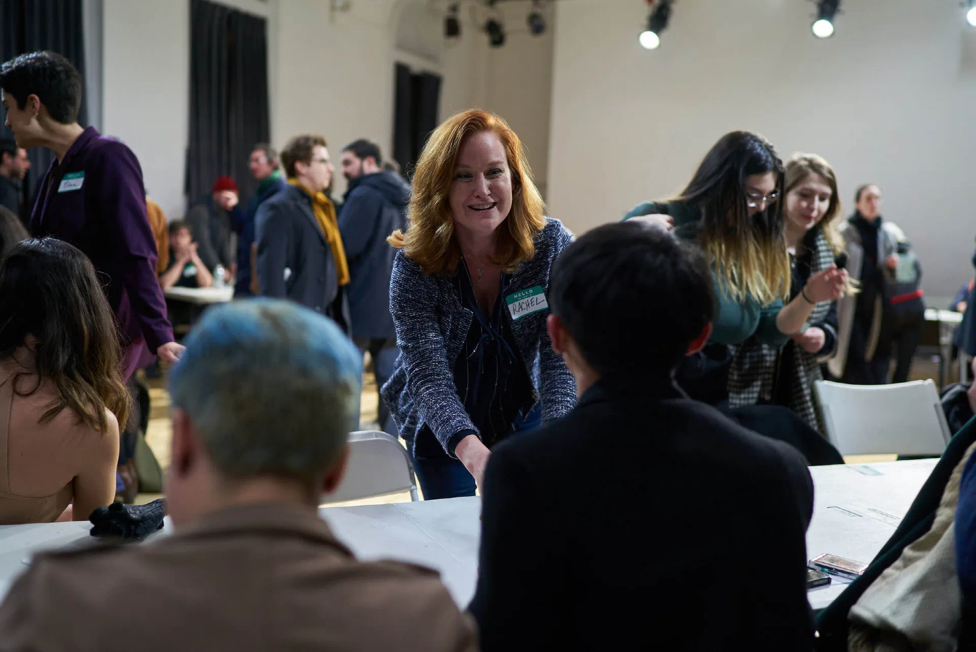 A woman speaks with participants seated around tables in a bright community hall.