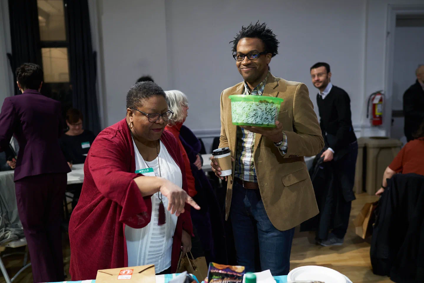 Two participants carry refreshments and materials through a crowded hall.