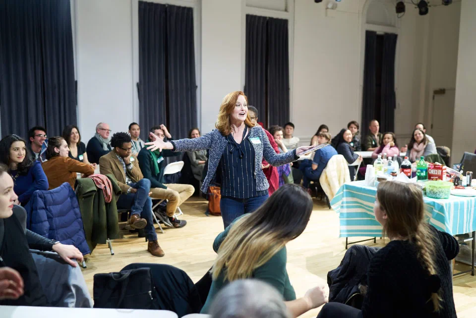 A performer addresses audience members seated around tables in a bright hall.