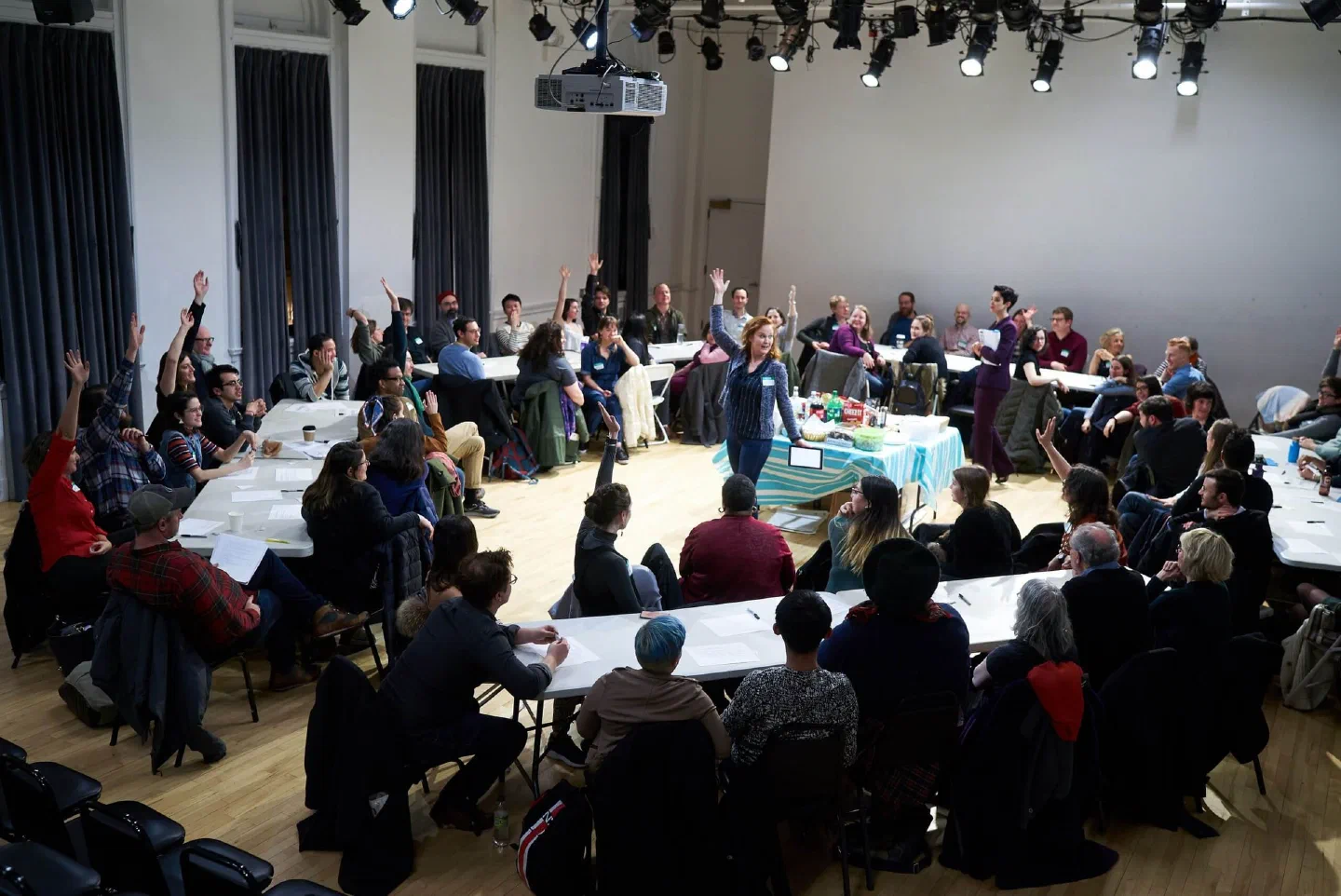 Audience members sit around tables as a performer addresses the room from the center.