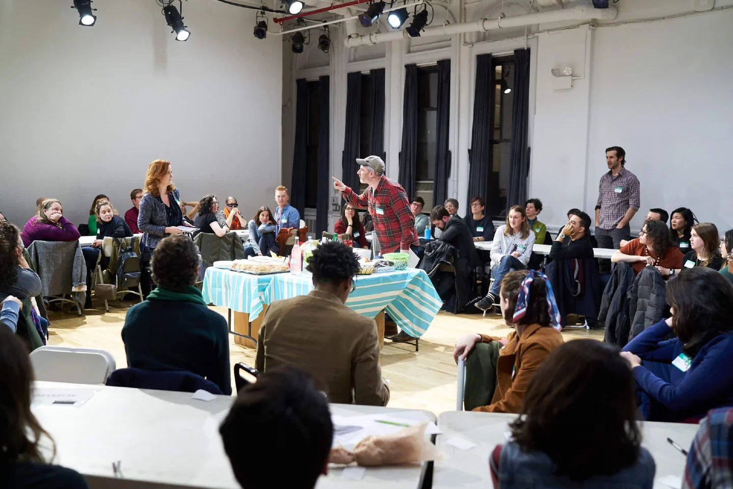 A performer stands among audience members seated at tables around the room.