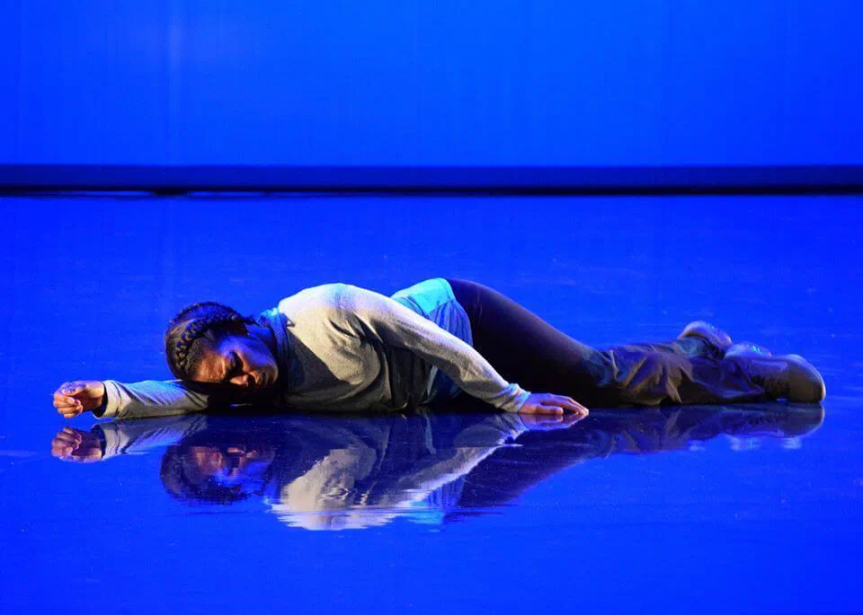 A performer lies curled on a reflective stage washed in vivid blue light.