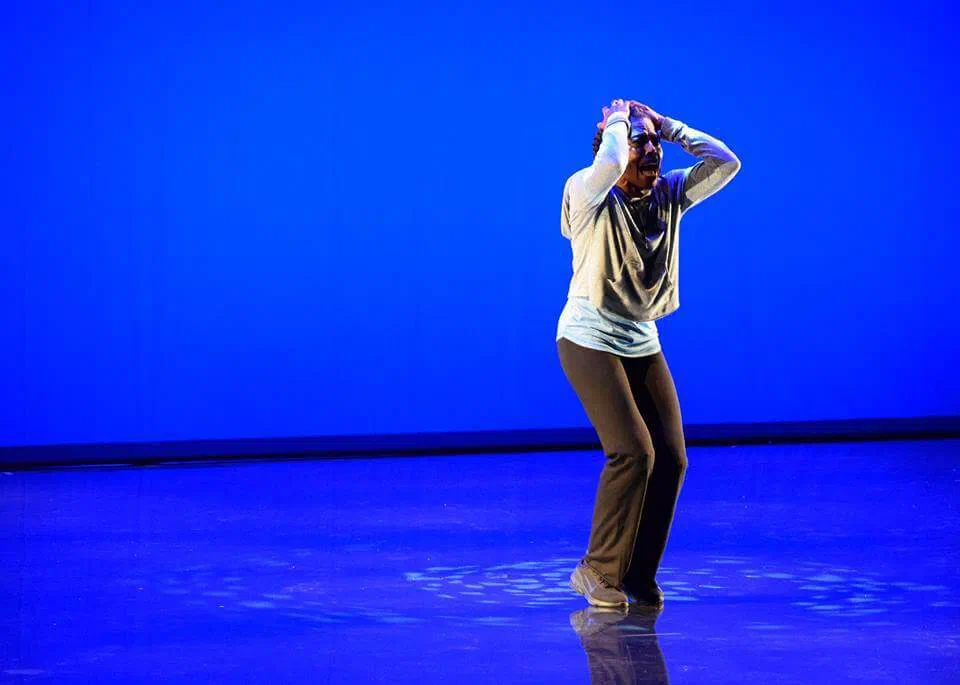 A lone performer holds both hands to their head against a vivid blue background.