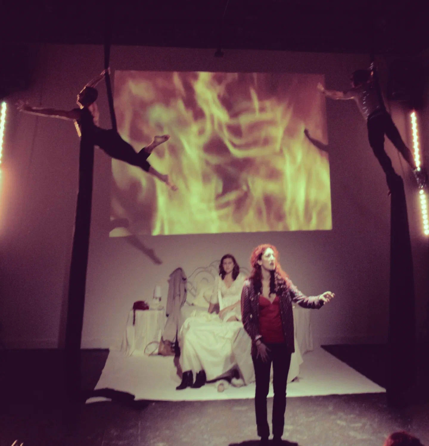 A woman gestures toward the audience as aerial performers hang above a bed and projected flames.