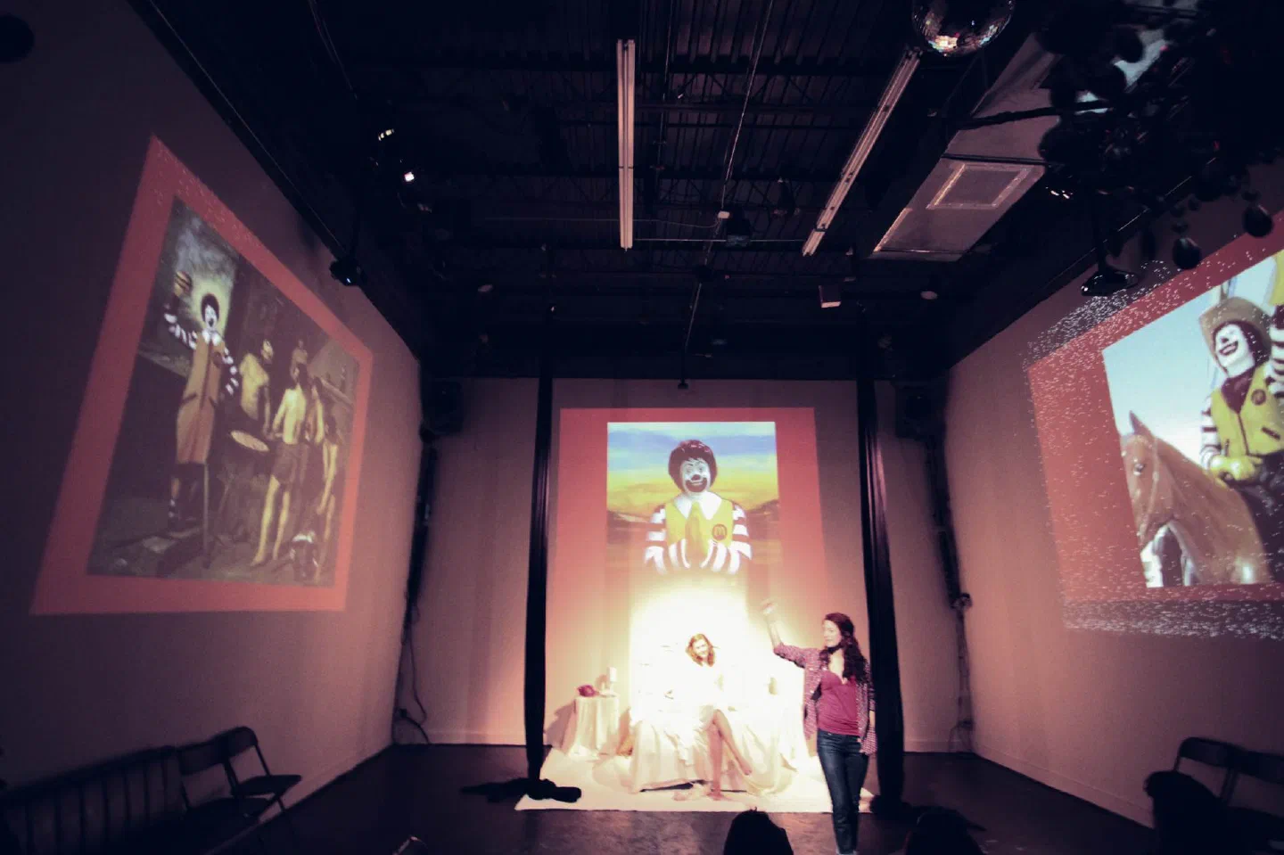 Two performers in white stand below projections of several costumed figures.