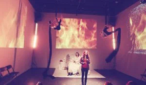 Two women perform beneath suspended figures and projections of flames.