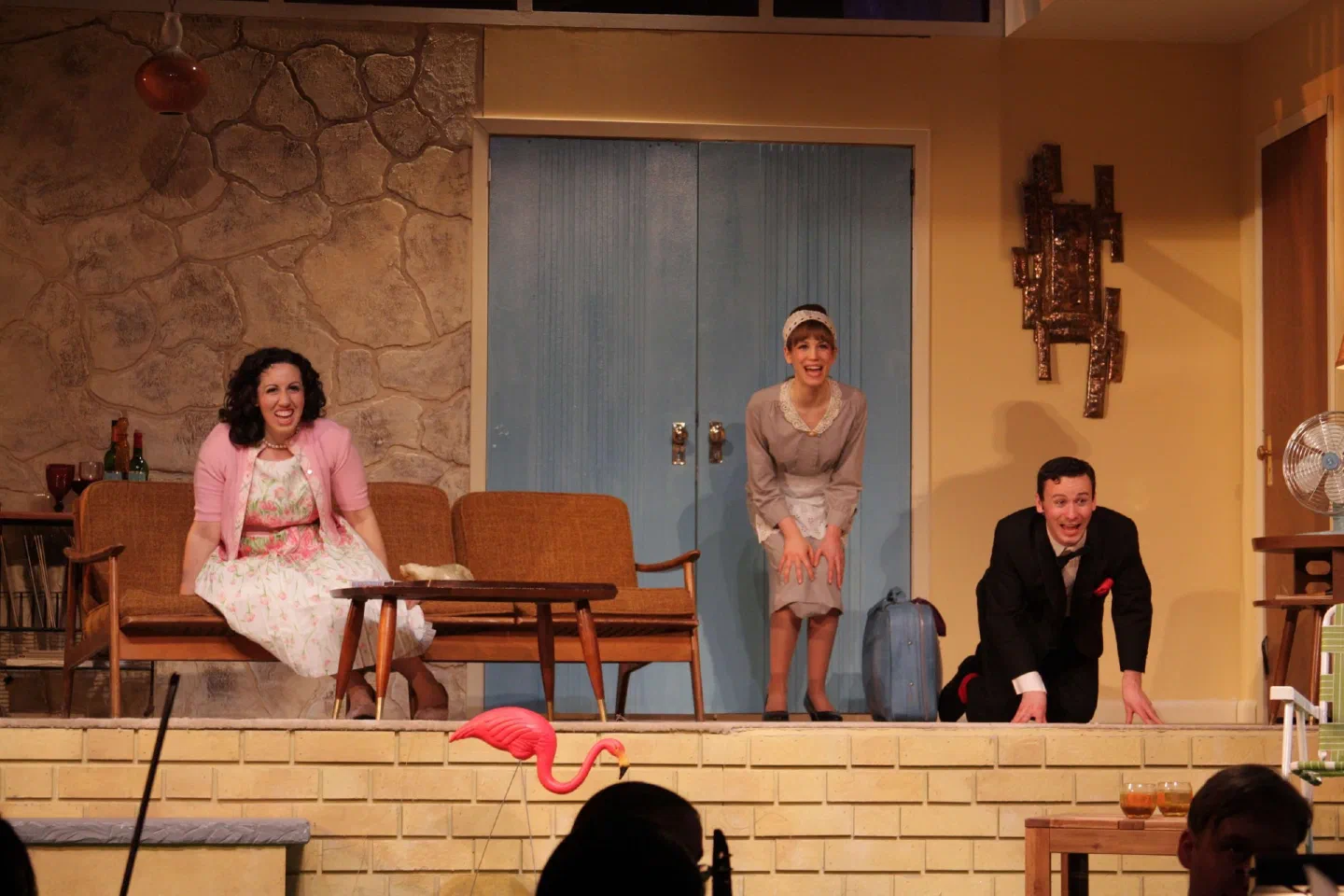 Three costumed performers sit and stand at the front of a domestic interior set.