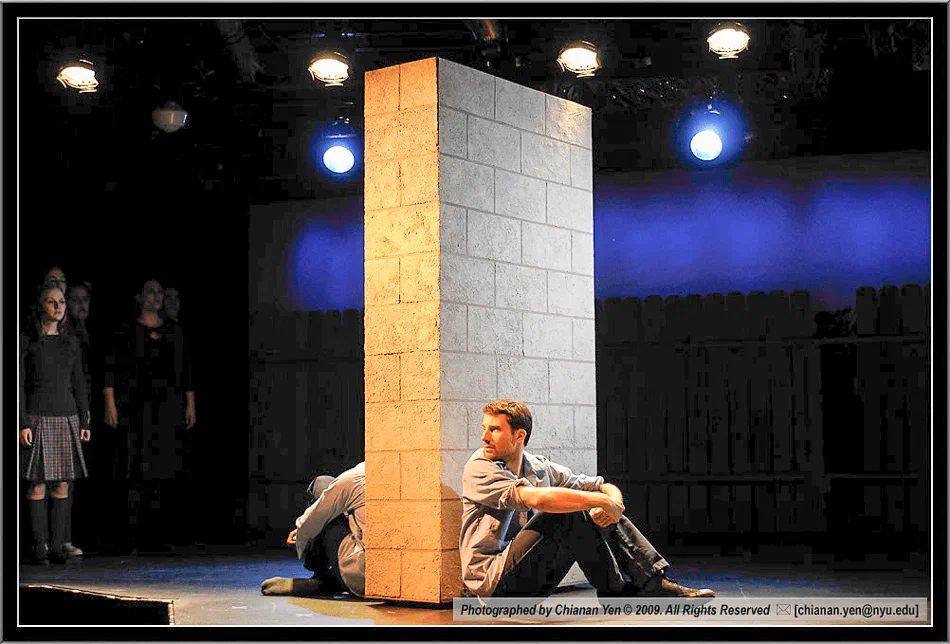 A performer sits beside a tall scenic wall under blue stage light.