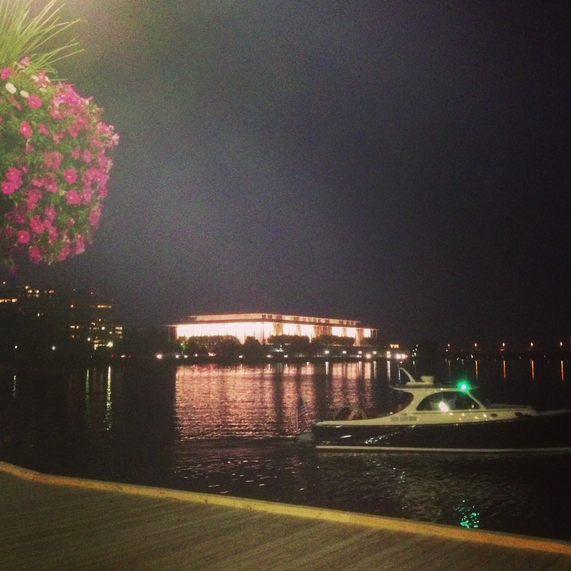 The Kennedy Center glows across the Potomac River at night, with a boat in the foreground.