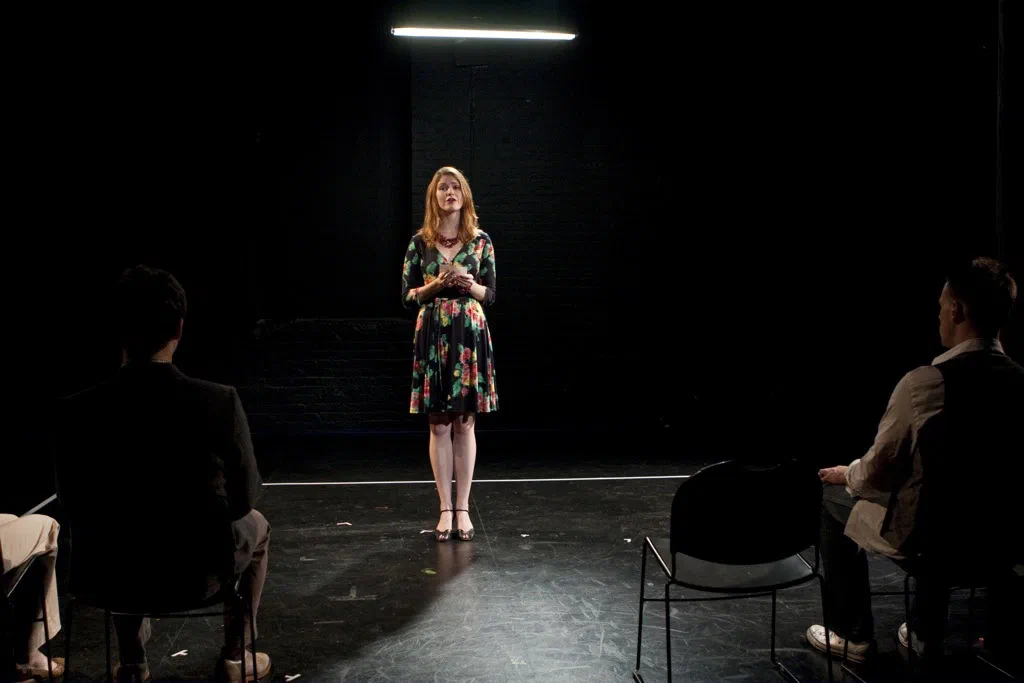 A woman in a floral dress stands beneath a fluorescent light facing seated audience members.