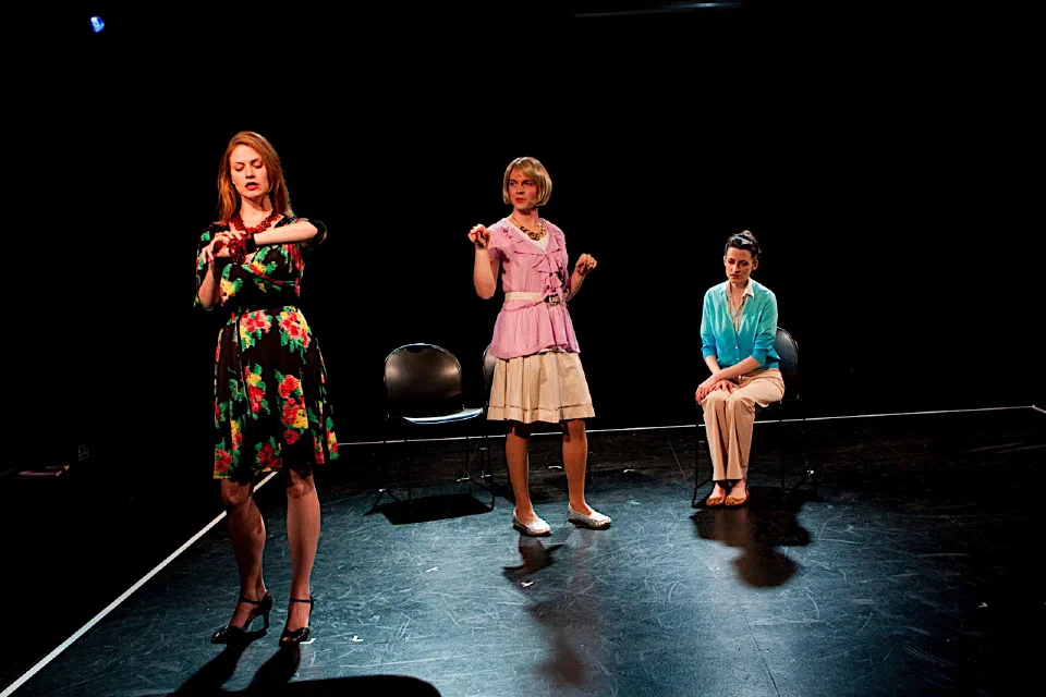 Three women in colorful dresses perform on a spare black stage.