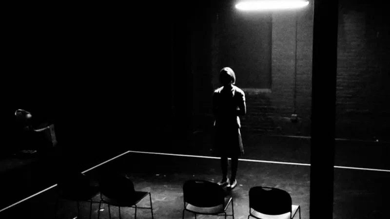 A lone performer stands in silhouette beneath a fluorescent light on a black stage.