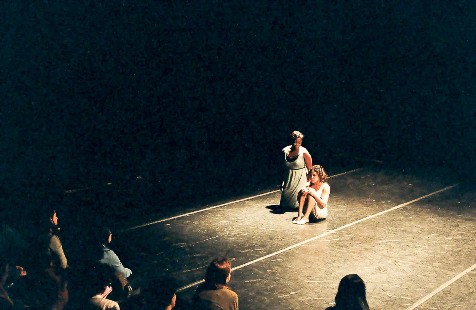 Two performers occupy a taped square of stage floor as audience members watch nearby.