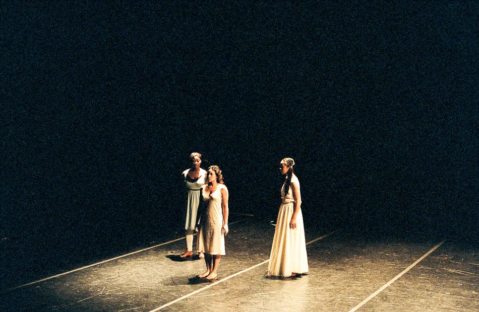 Three performers in pale costumes stand together in a pool of stage light.