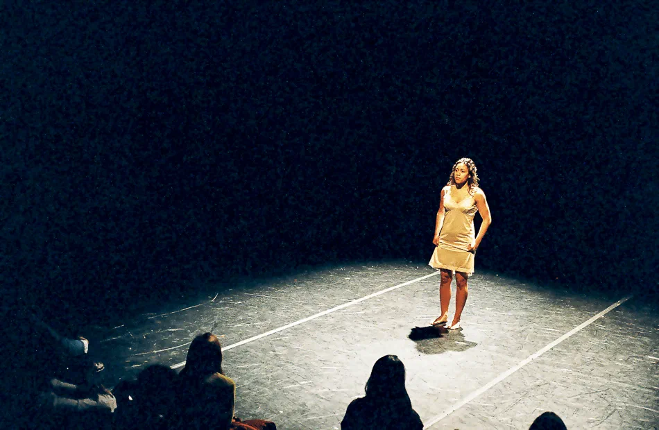 A lone woman stands in a pool of light within a taped square on the stage floor.