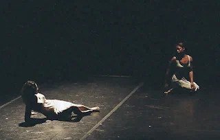 One performer reclines while another kneels nearby on a dimly lit stage.