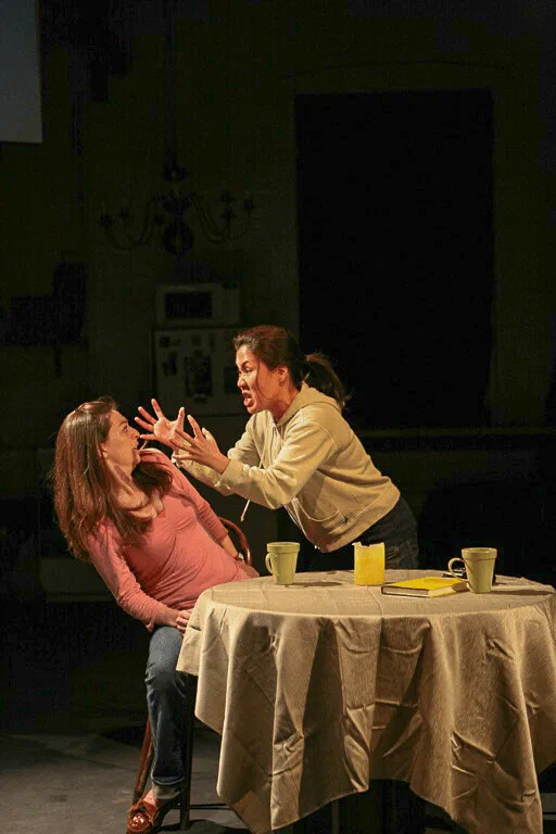 One woman gestures toward another who sits with her head bowed at a small table.