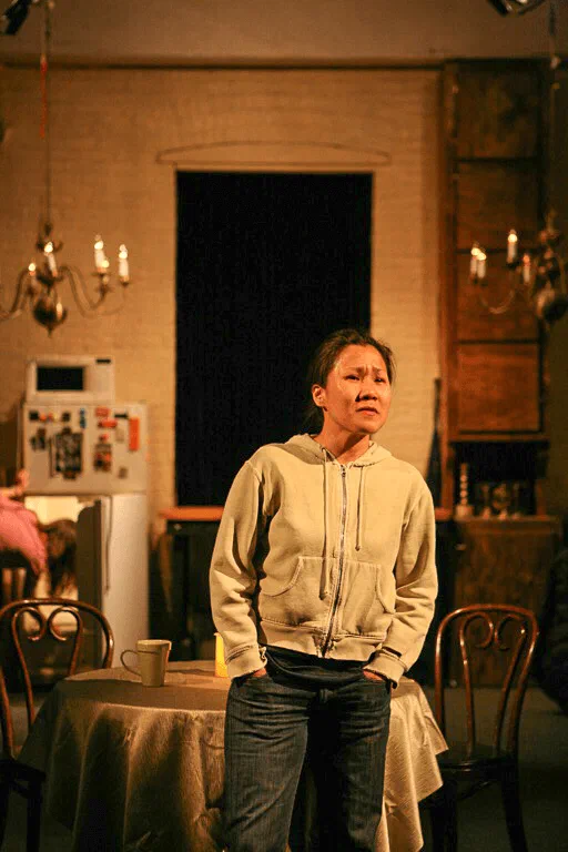 A woman stands beside a round table at the front of a domestic interior.