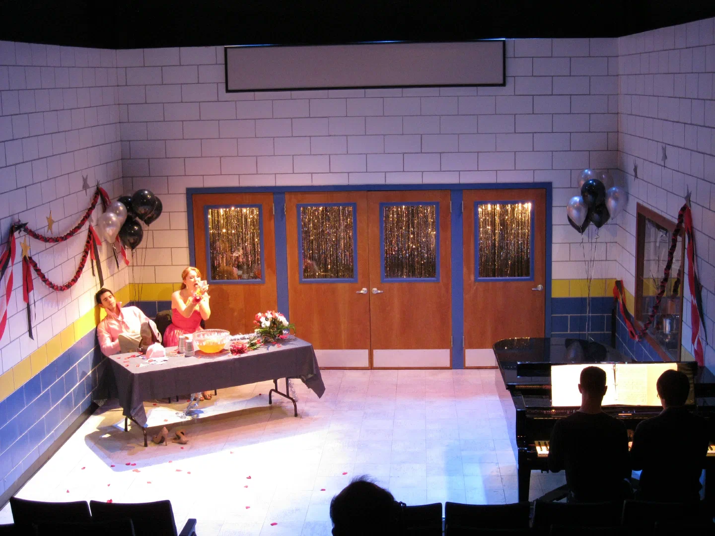 Two women sit at a table on a brightly lit set decorated with streamers and balloons.