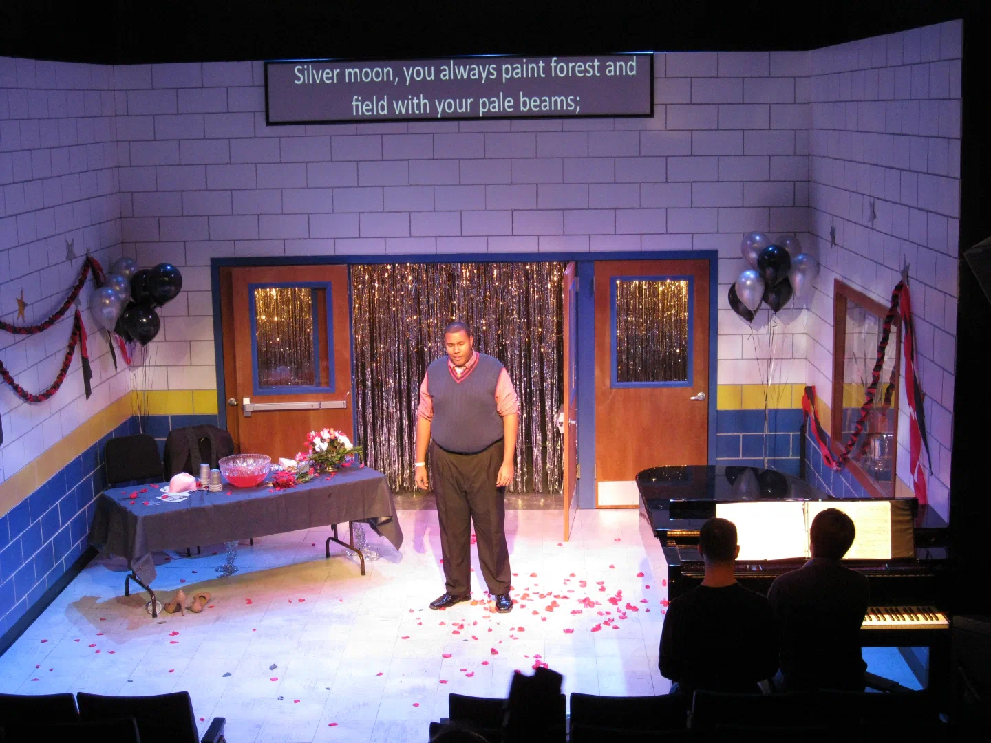 A lone man stands amid red flower petals beneath a screen of projected text.