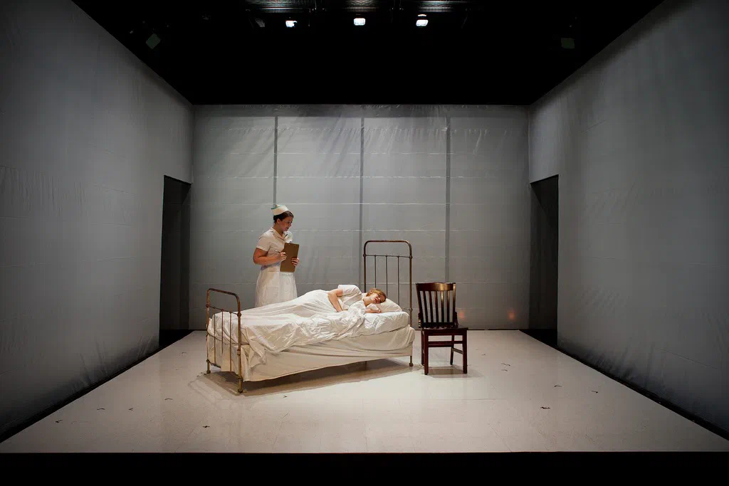 A uniformed figure stands beside a patient lying in a metal-framed bed on a stark white set.