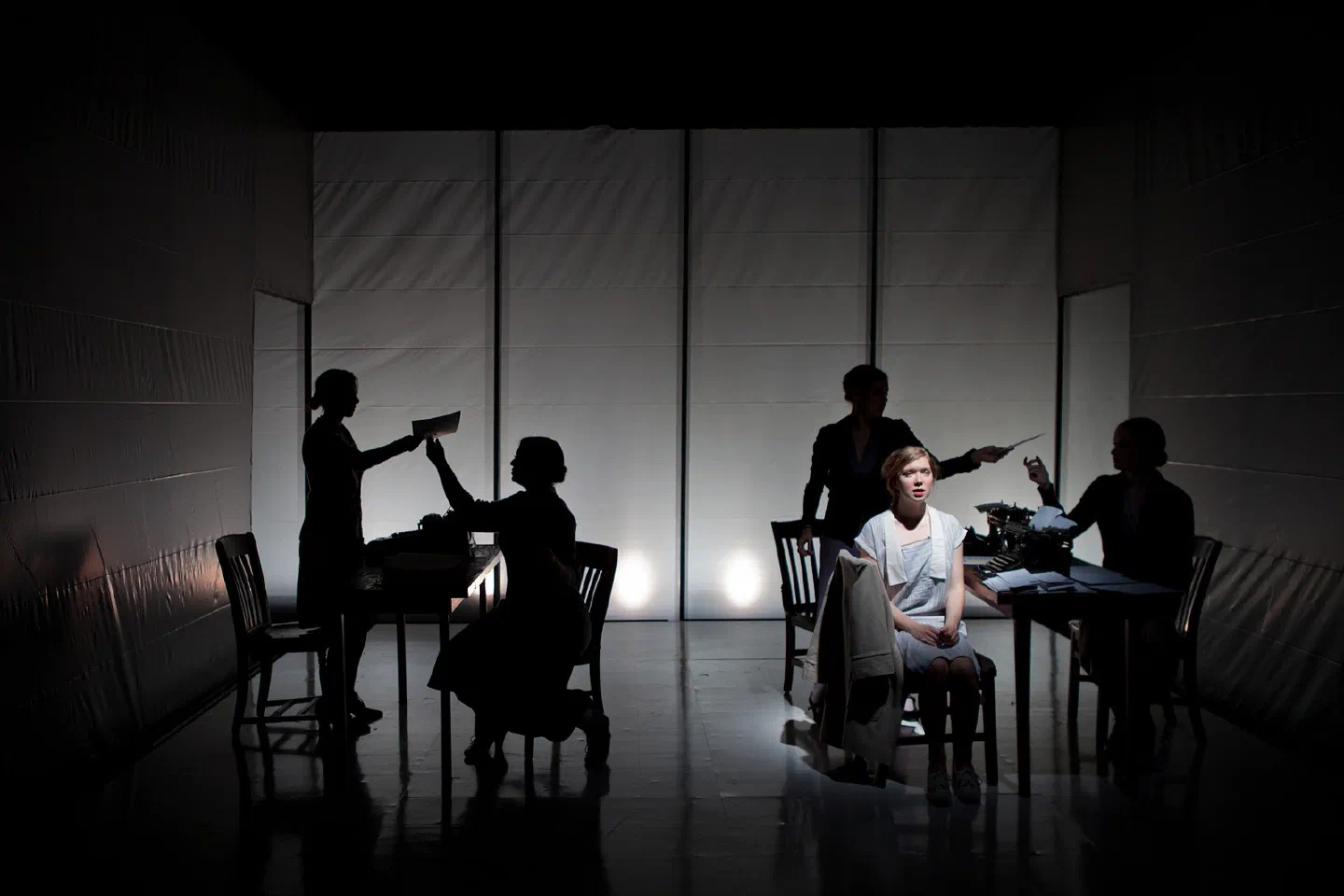 A seated woman is encircled by silhouetted figures holding papers and bottles.