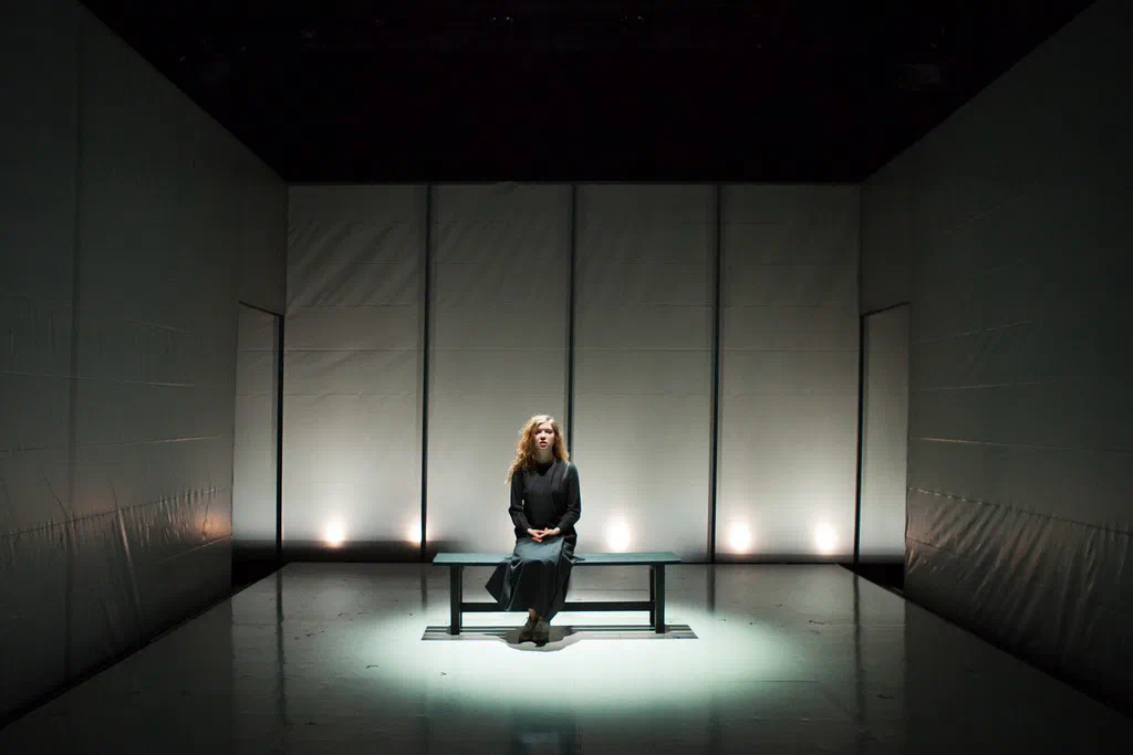 A lone woman sits on a bench in a circular pool of light within a white room.