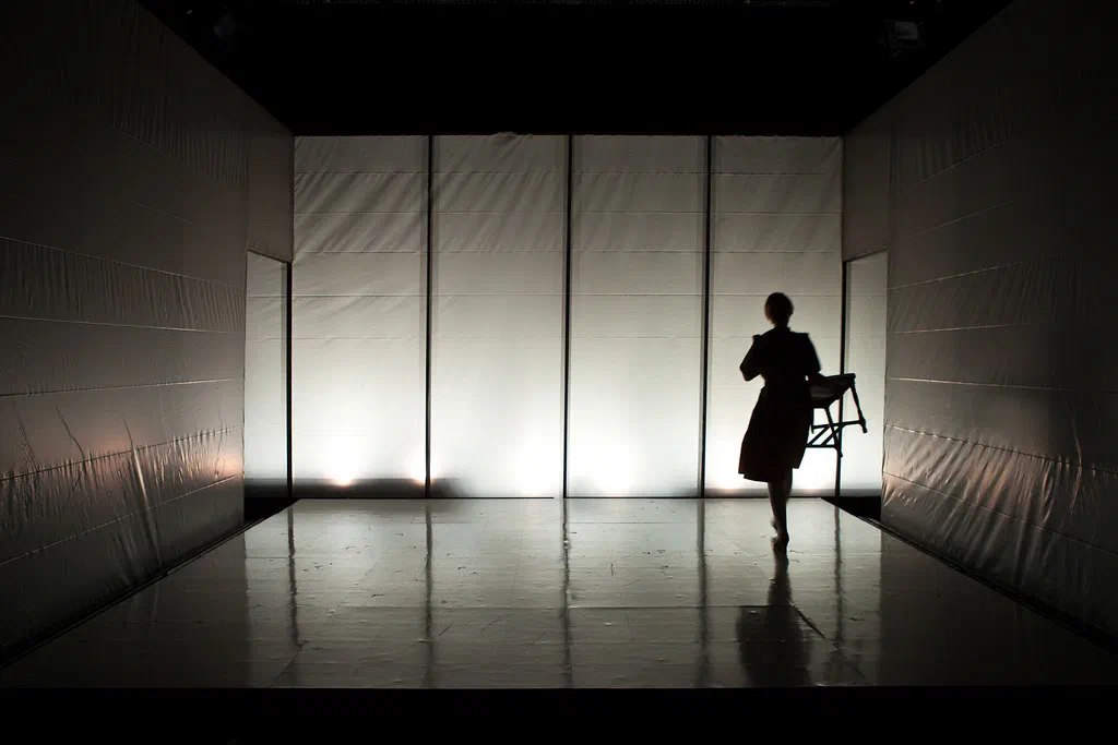 A silhouetted woman crosses an empty white room carrying a handbag.