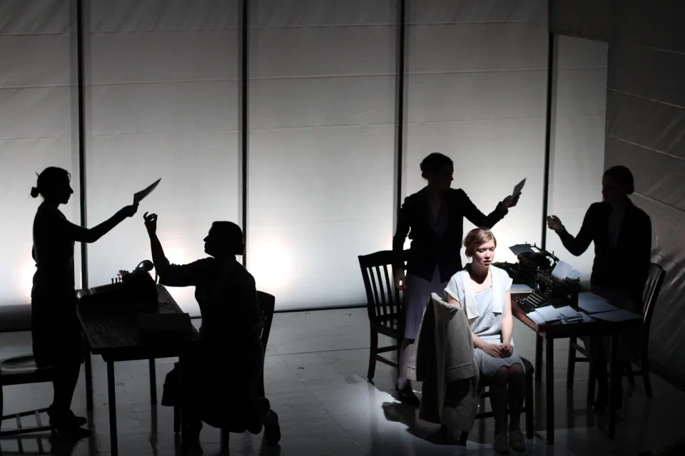 A seated woman is surrounded by sharply silhouetted figures at a table.