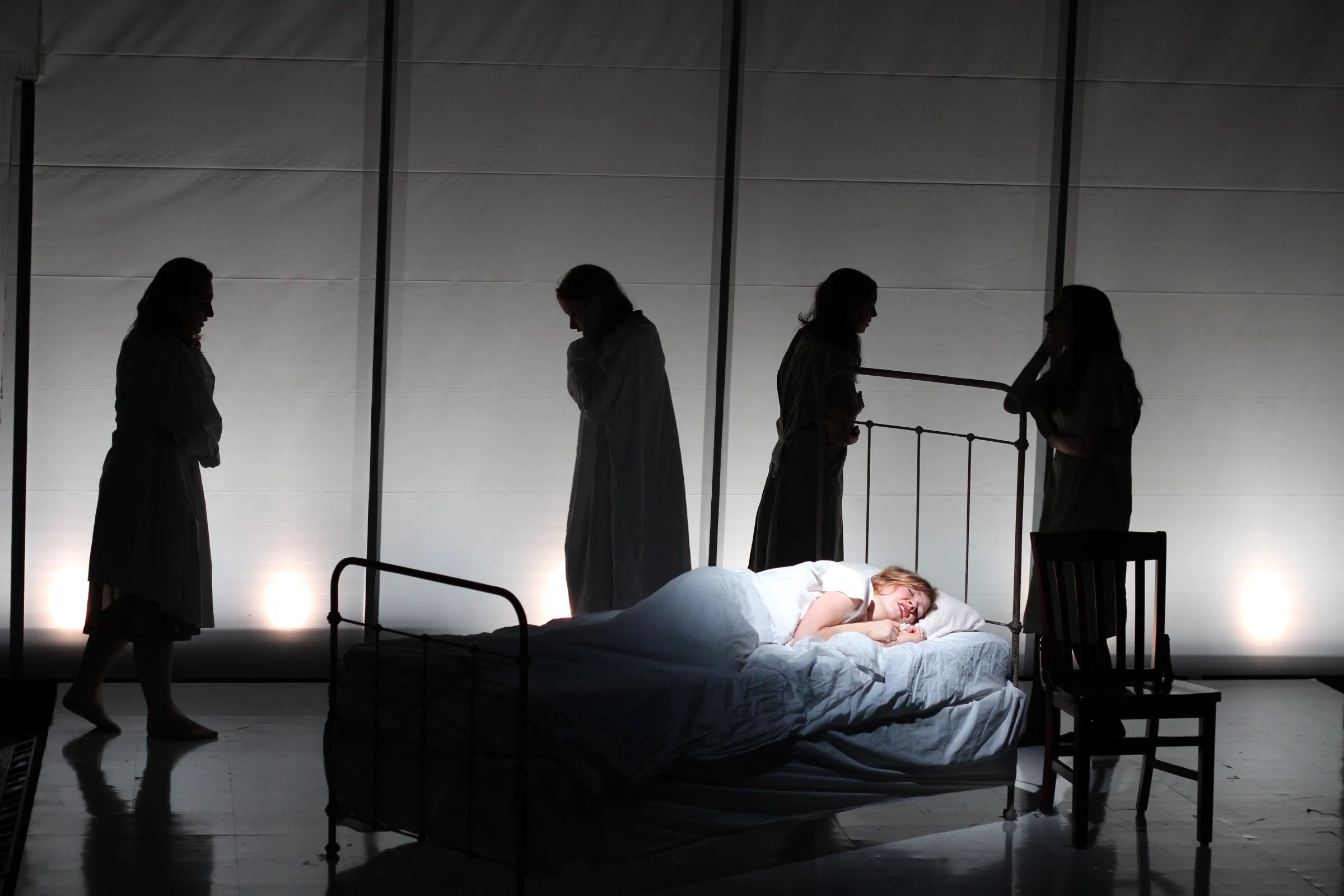 Shadowed figures gather around a woman lying in a bed on a stark white set.