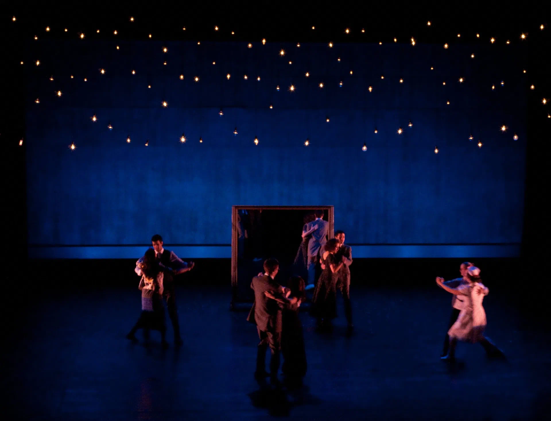 An ensemble performs beneath rows of small lights on a deep blue stage.