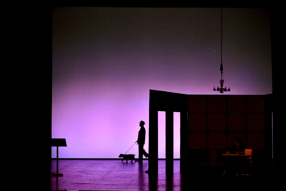 A silhouetted performer crosses a geometric set washed in purple light.