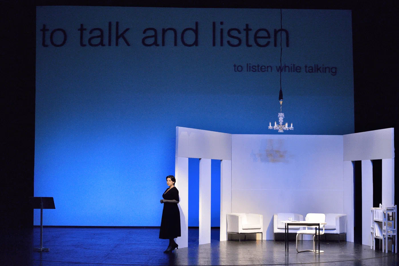 A woman stands in a white room beneath the projected words to talk and listen.
