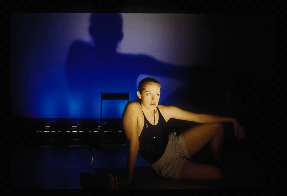 A woman sits on the floor beneath an enlarged shadow projected on the wall behind her.