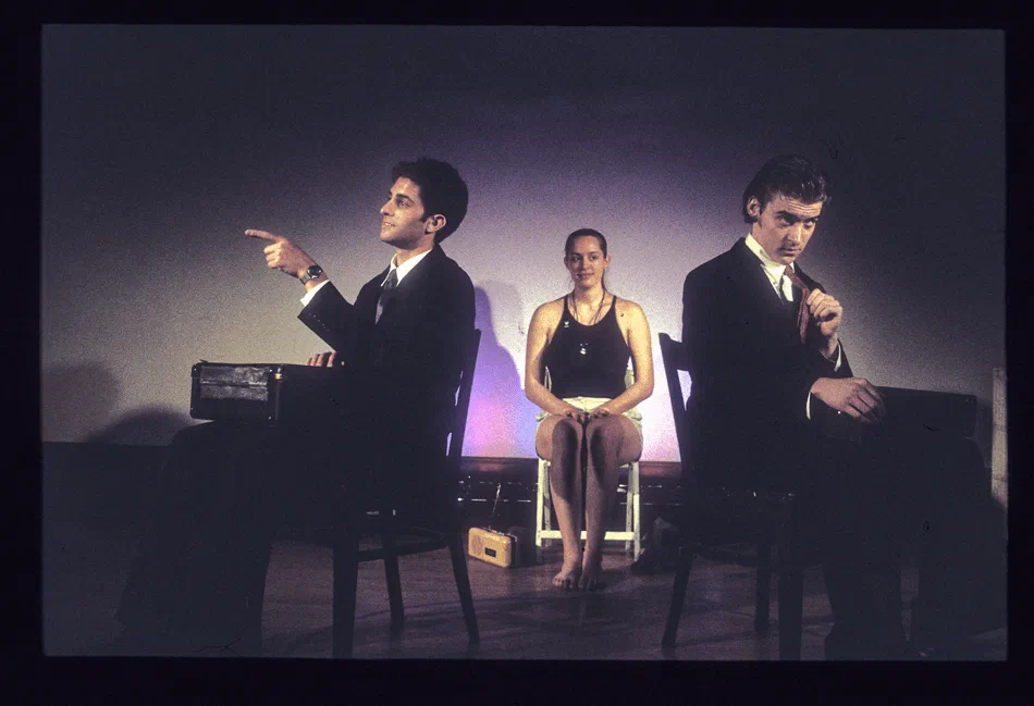 Two suited men point in opposite directions while a woman sits between them.