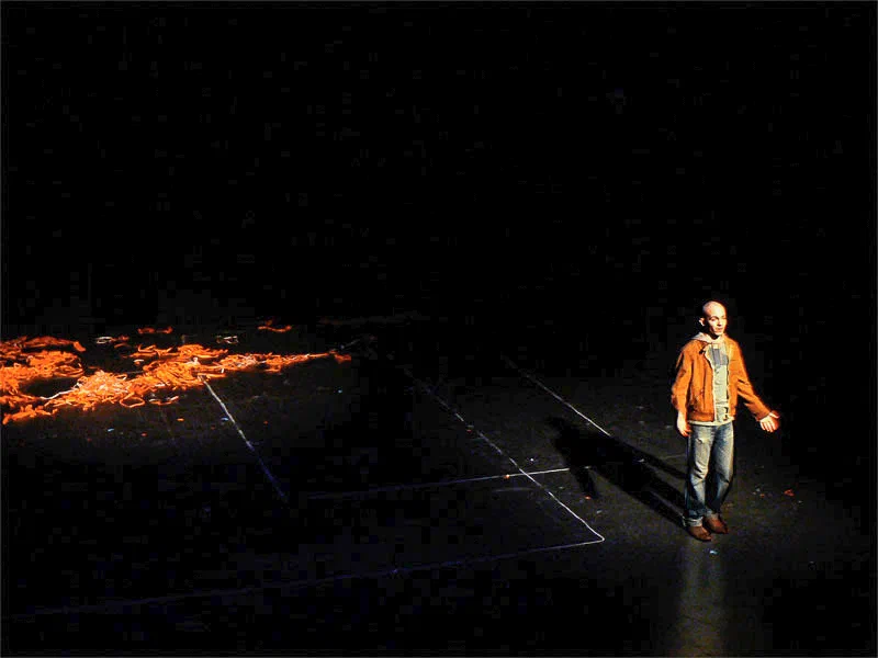 A lone man in an orange jacket stands at the edge of a wide dark stage.