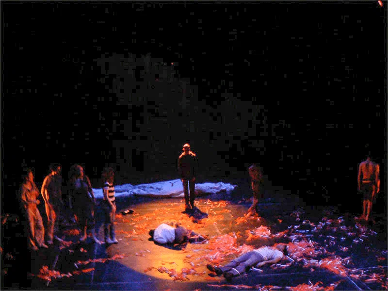 An ensemble surrounds two figures lying amid feathers and debris under red light.