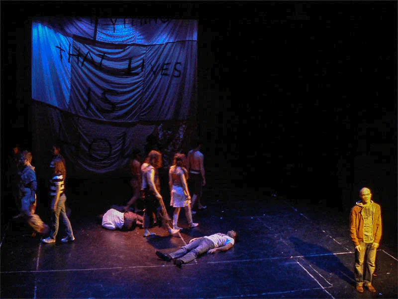 Performers gather around prone figures beneath a large hanging fabric banner.