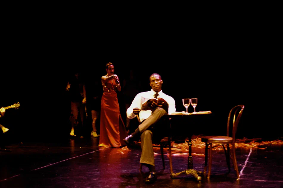 A man sits beside a small table and wine glasses while a woman stands behind him.