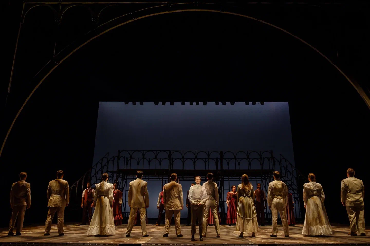 A large costumed ensemble faces a bright backdrop at the rear of the stage.