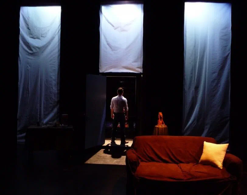 A lone performer stands between hanging white fabric panels and a red couch.