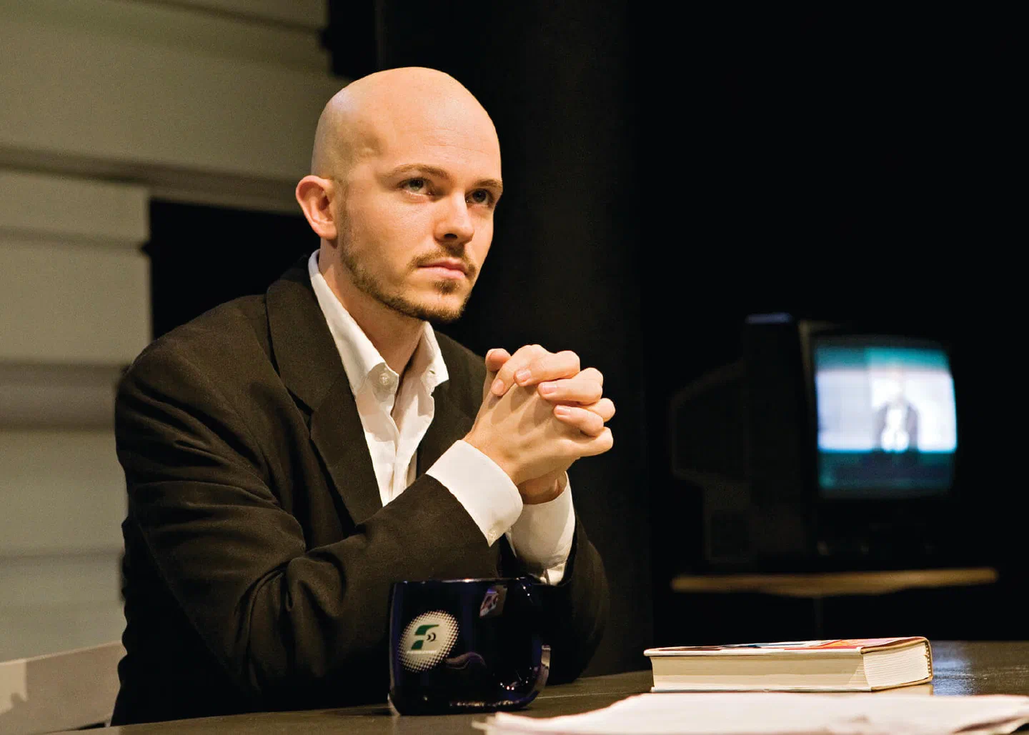 A bald man in a dark suit sits with clasped hands at a desk beside a television.