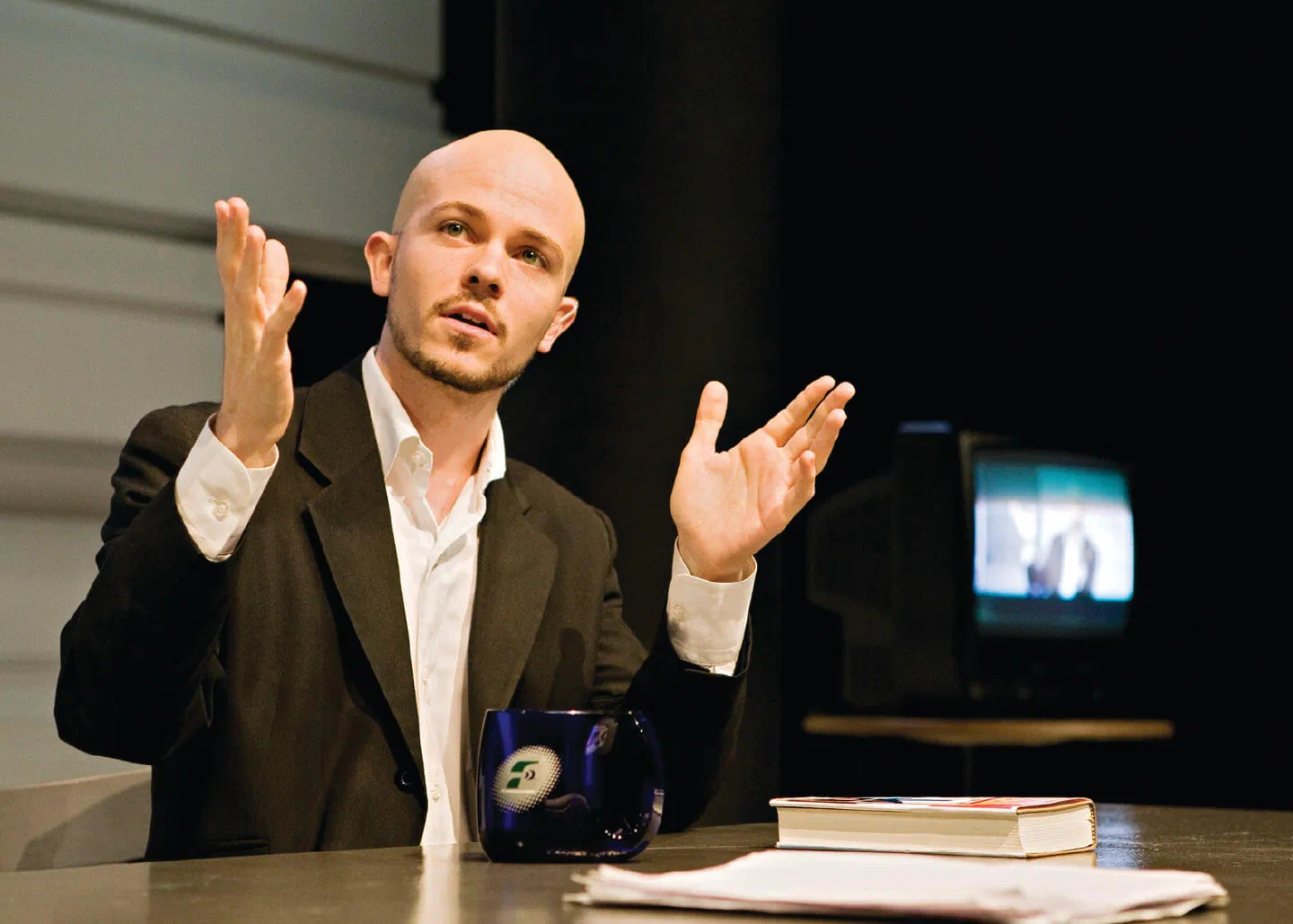 A seated man gestures with both hands above a desk holding books and a dark mug.